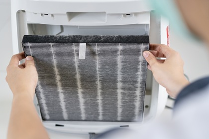Someone placing a replacement filter into a portable air cleaner device. The filter has a folded structure like a HEPA filter and dark material like a carbon filter.