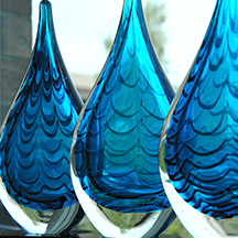 Three blown glass art pieces are placed one in front of the other, each slightly to the right so the perspective overlaps. Each piece is similar in shape and design but unique. They have a teardrop shape, with clear glass for the exterior and blue rippled glass for the interior. The thickness of the clear glass increases from top to bottom, and takes up about 20% of the height at the base. The rippled blue interior looks like a drop of water within the clear exterior.
