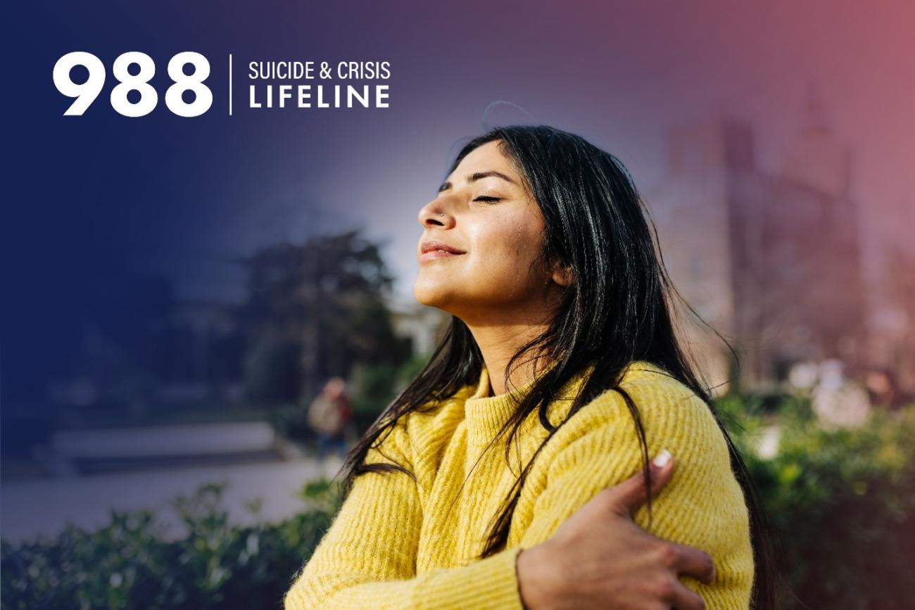 988 Suicide and Crisis Lifeline at the top left with woman in a yellow colored sweater with black hair and tan colored skin wrapping her arms around her body.