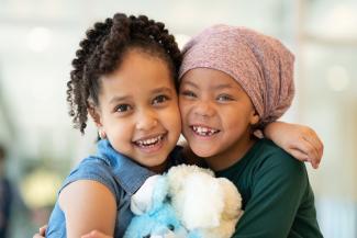 Two children look at the camera while hugging. One had curly black hair and one has a bandana wrapped around her head.